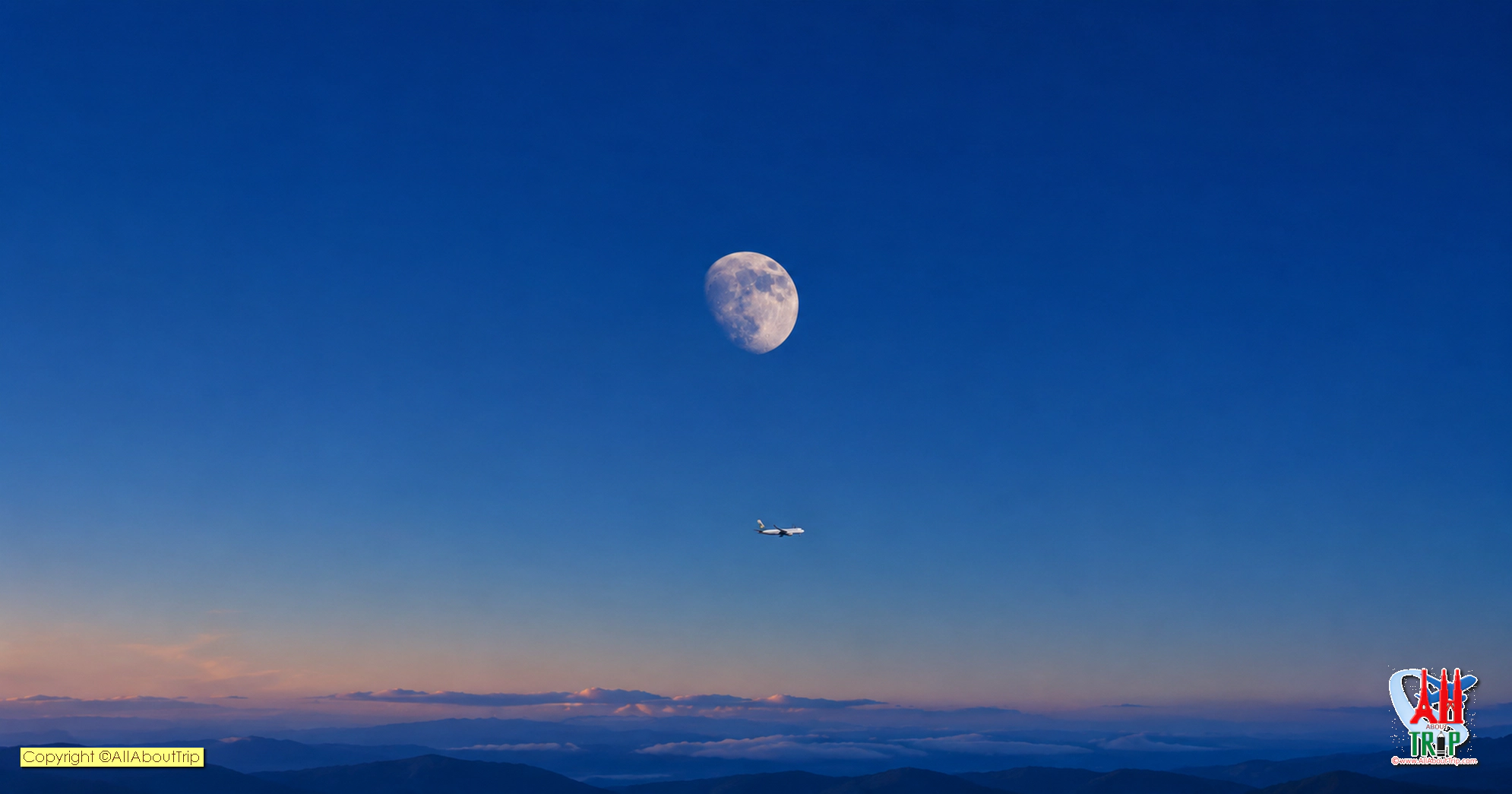 A passenger airplane beneath the moon above distant mountains at twilight.